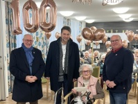 100. Geburtstag Frau Korthues (15. Januar) Drei Männer neben einer Seniorin im Rollstuhl mit goldenen Ballons und der Aufschrift "Happy Birthday", vor einem Raum mit sitzenden Menschen