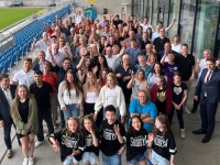 Eine Gruppe von etwa 100 Menschen posiert für ein Gruppenfoto in der Zuschauertribühne eines Stadions. 