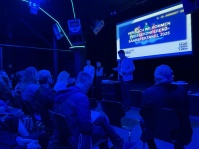 Ein Mann mit Mikrofon steht vor einem sitzenden Publikum und hält in einem Raum mit besonderem Licht eine Rede