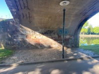 Ein Weg führt unter einer Brücke her, im Hintergrund ein Fluss