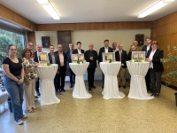 Eine Gruppe Menschen in feierlicher Kleidung steht nebeneinander an geschmückten Stehtischen in einem Saal.