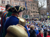 Ein Mann im blauen Kostüm mit Dreispitz steht auf der Bühne vor dem Rathaus und blickt auf eine große Menschenmenge.