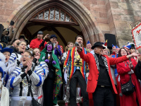 Eine Gruppe kostümierter Menschen steht auf den Treppen vor dem Rathauseingang, während ein Mann im bunten Kostüm in ein Mikrofon spricht.