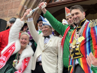 Mehrere kostümierte Personen stehen vor dem Rathauseingang und singen oder rufen gemeinsam in Richtung Publikum.