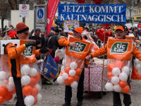 Mehrere Männer in orangefarbenen Tic-Tac-Kostümen stehen mit einem Schild „Jetzt kommt kaltnaggisch“ vor einer großen Menschenmenge.