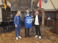 Drei Personen stehen in einem Saal. Die Person in der Mitte hält ein blaues Fußballtrikot mit der Aufschrift „Conradt“.